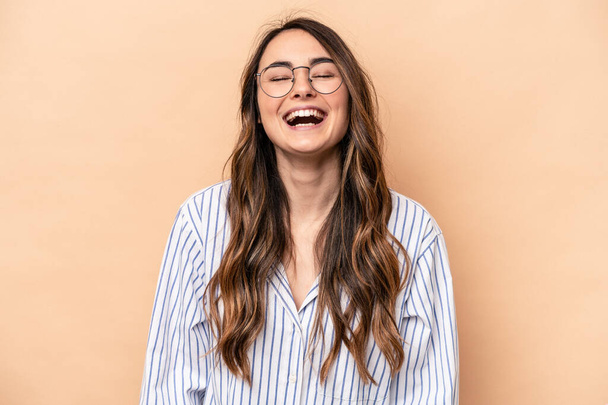 Young caucasian woman isolated on beige background relaxed and happy laughing, neck stretched showing teeth. - Photo, Image