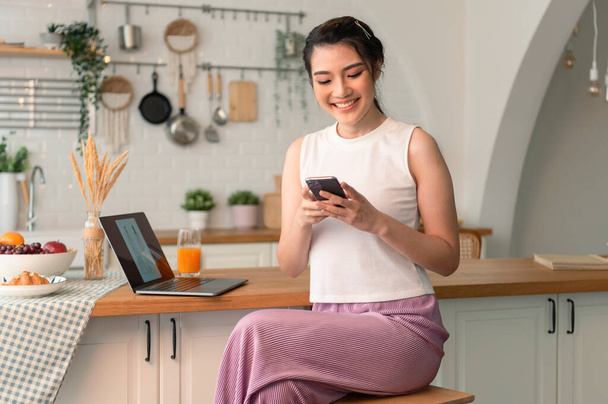 Smiling young asian woman using mobile phone while sitting in kitchen room at home with laptop computer. - Photo, Image