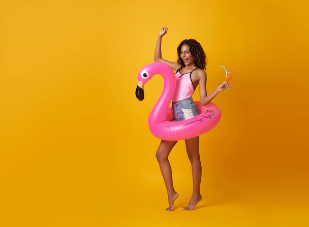 Happy woman holding orange juice with flamingo rubber ring on yellow copy space background. woman relaxing on beach in summer holiday. - Foto, Imagem