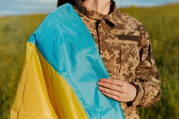 Female soldier dressed in military uniform wrapped in ukrainian flag symbol of independence. Armed Forces of Ukraine. Woman serving in ukrainian army. - 写真・画像