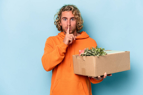 Young caucasian man making a move while picking up a box full of things isolated on blue background keeping a secret or asking for silence. - Foto, immagini