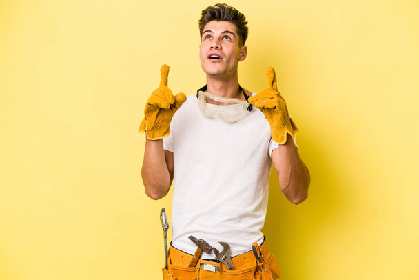 Young electrician caucasian man isolated on yellow background pointing upside with opened mouth. - Fotoğraf, Görsel