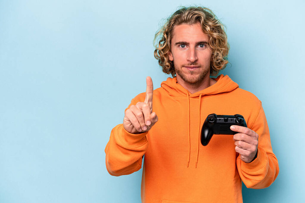 Young caucasian man playing with a video game controller isolated on blue background showing number one with finger. - Foto, imagen