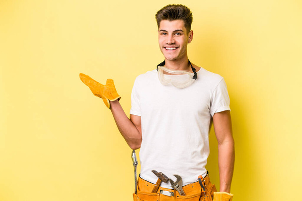 Молодий електрик кавказький чоловік ізольований на жовтому тлі, показуючи простір для копіювання на долоні і тримаючи іншу руку на талії
. - Фото, зображення