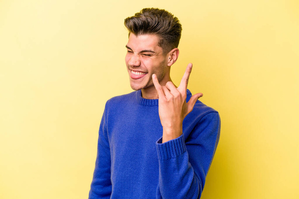 Young caucasian man isolated on yellow background showing rock gesture with fingers - Photo, Image