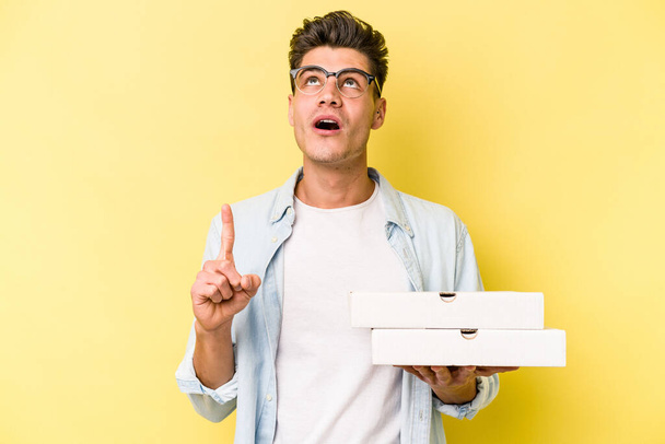 Young caucasian man holding pizzas isolated on yellow background pointing upside with opened mouth. - Zdjęcie, obraz