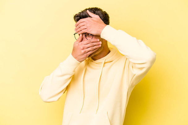 Νεαρός καυκάσιος άνδρας απομονωμένος σε κίτρινο φόντο ανοιγοκλείνει τα μάτια του στην κάμερα μέσα από δάχτυλα, ντροπιασμένος καλύπτοντας το πρόσωπο. - Φωτογραφία, εικόνα