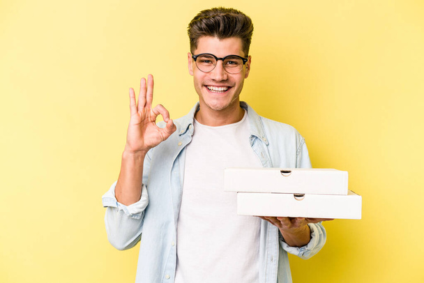Young caucasian man holding pizzas isolated on yellow background cheerful and confident showing ok gesture. - Foto, afbeelding