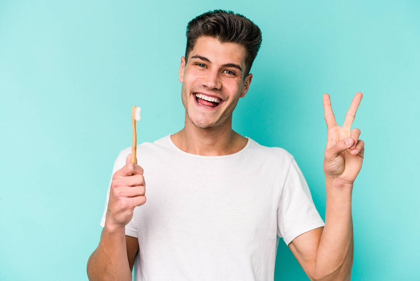 Young caucasian man brushing teeth isolated on white background joyful and carefree showing a peace symbol with fingers. - Fotografie, Obrázek