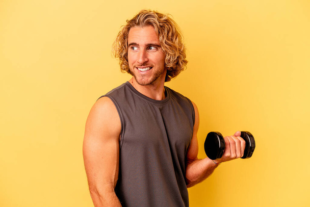 Jonge sportman maakt gewichtheffen geïsoleerd op gele achtergrond kijkt opzij glimlachend, vrolijk en aangenaam. - Foto, afbeelding