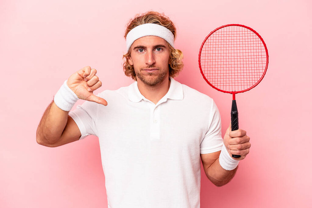 Junger kaukasischer Mann, der isoliert auf rosa Hintergrund Badminton spielt und eine ablehnende Geste zeigt, Daumen nach unten. Konzept der Uneinigkeit. - Foto, Bild