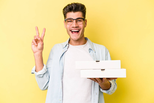 Young caucasian man holding pizzas isolated on yellow background joyful and carefree showing a peace symbol with fingers. - Foto, afbeelding