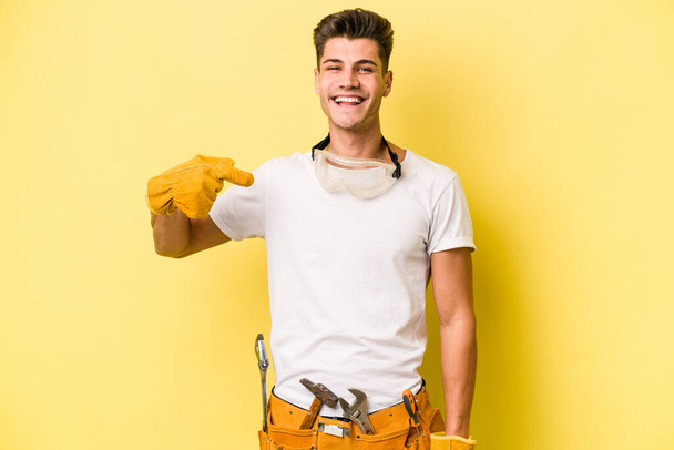 Jeune électricien caucasien isolé sur fond jaune personne pointant à la main vers un espace de copie chemise, fier et confiant - Photo, image
