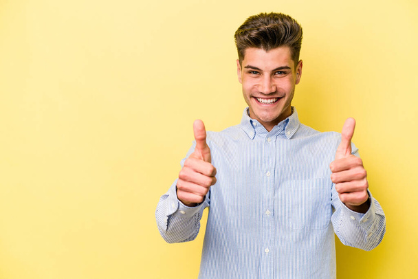 Fiatal fehér férfi elszigetelt sárga háttér mosolyog és felemeli hüvelykujját - Fotó, kép