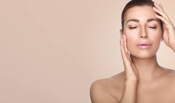 Beautiful middle-aged woman with healthy flawless skin posing with hands to cheeks and eyes closed over a beige background with copyspace in a skincare concept - Photo, image
