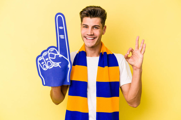Jonge sportfan man geïsoleerd op gele achtergrond vrolijk en vol vertrouwen tonen ok gebaar. - Foto, afbeelding