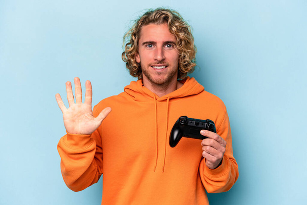 Mavi arka planda izole edilmiş bir bilgisayar oyunu kontrolörüyle oynayan beyaz bir adam beş numarayı parmaklarıyla gülümsüyor.. - Fotoğraf, Görsel