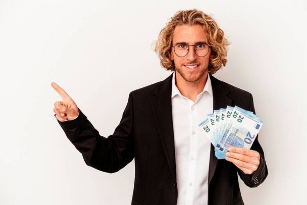 Молодой кавказский бизнесмен держит банкноты на белом фоне, улыбаясь и указывая в сторону, показывая что-то на пустом месте. - Фото, изображение