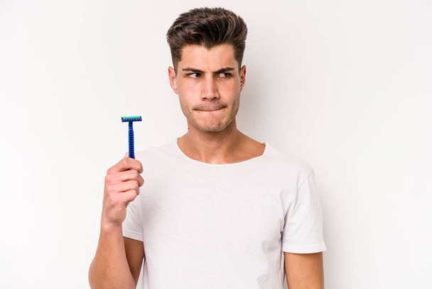 Young caucasian man shaving his beard isolated on white background confused, feels doubtful and unsure. - Foto, Imagen