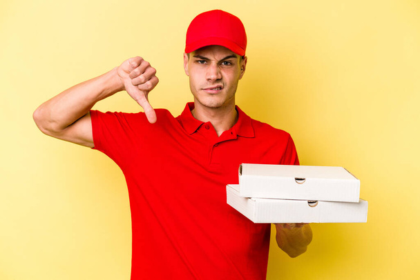 Młody mężczyzna trzymający pizzę na żółtym tle, wykazujący niechęć do gestów, opuszczone kciuki. Koncepcja sporu. - Zdjęcie, obraz