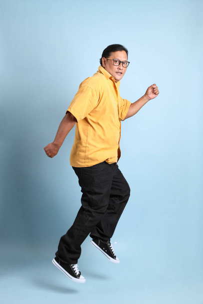 The adult Asian man with yellow shirt jumping on the blue background. - Fotó, kép