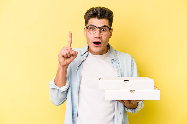 Young caucasian man holding pizzas isolated on yellow background having an idea, inspiration concept. - Fotoğraf, Görsel