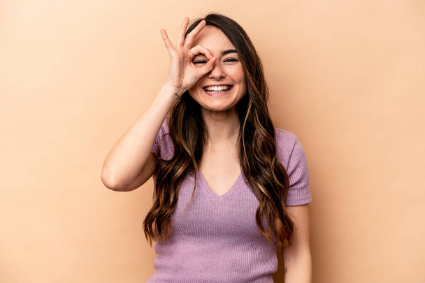 Young caucasian woman isolated on beige background excited keeping ok gesture on eye. - Foto, Imagem