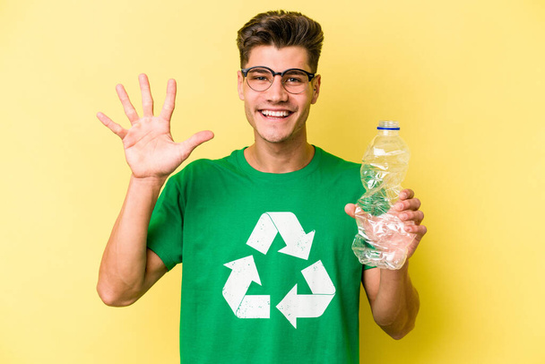 Sarı arka planda izole edilmiş geri dönüşüm için elinde plastik şişesi olan beyaz bir adam gülümseyerek beş numarayı parmaklarıyla gösteriyor.. - Fotoğraf, Görsel