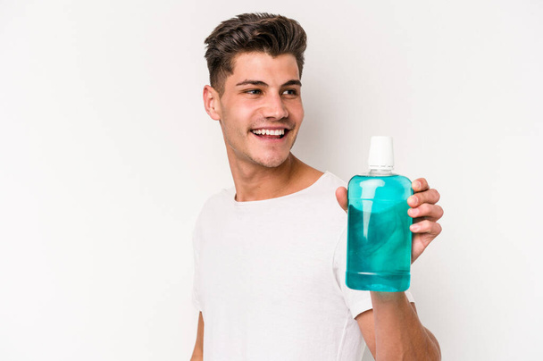 Young caucasian man holding mouthwash isolated on white background looks aside smiling, cheerful and pleasant. - Foto, Imagen
