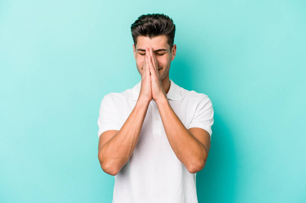 Young caucasian man isolated on blue background holding hands in pray near mouth, feels confident. - Photo, Image