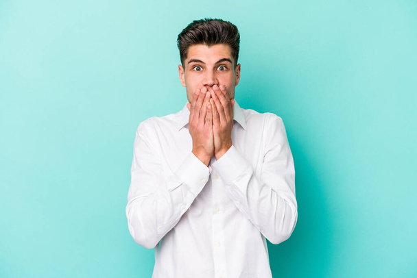 Young caucasian man isolated on blue background shocked covering mouth with hands. - Zdjęcie, obraz