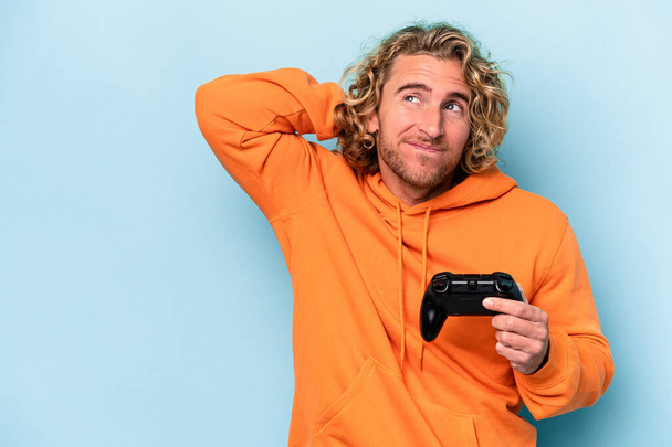 Junger kaukasischer Mann spielt mit einem Videospielcontroller isoliert auf blauem Hintergrund, berührt den Hinterkopf, denkt nach und trifft eine Entscheidung. - Foto, Bild
