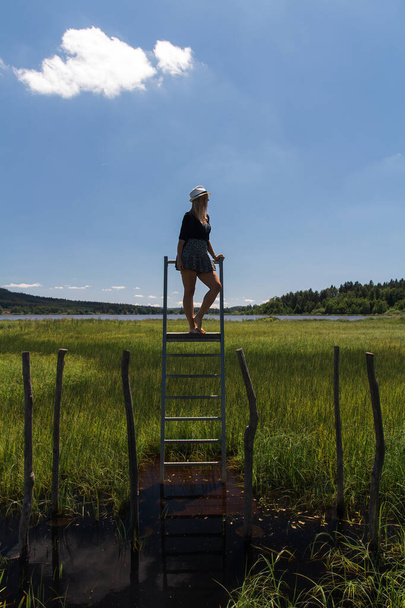 Junge Frau mit Hut steht auf Leiter auf Lehrpfad Olsina von der Seite. Böhmerwald, tschechische Sommerlandschaft - Foto, Bild