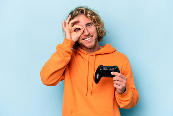 Νεαρός καυκάσιος άνδρας παίζει με ένα χειριστήριο βιντεοπαιχνιδιών απομονωμένο σε μπλε φόντο ενθουσιασμένος κρατώντας εντάξει χειρονομία στο μάτι. - Φωτογραφία, εικόνα