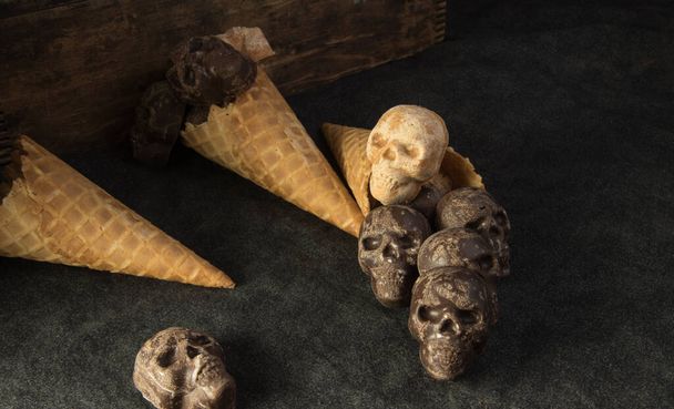 Kísérteties, csokis és mogyoróvajas cukorkás koponyák ömlenek ki gofris tölcsérből sötét háttérrel. Halloween buli.. - Fotó, kép
