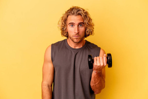 young sport man making weightlifting isolated on yellow background shrugs shoulders and open eyes confused. - Foto, imagen