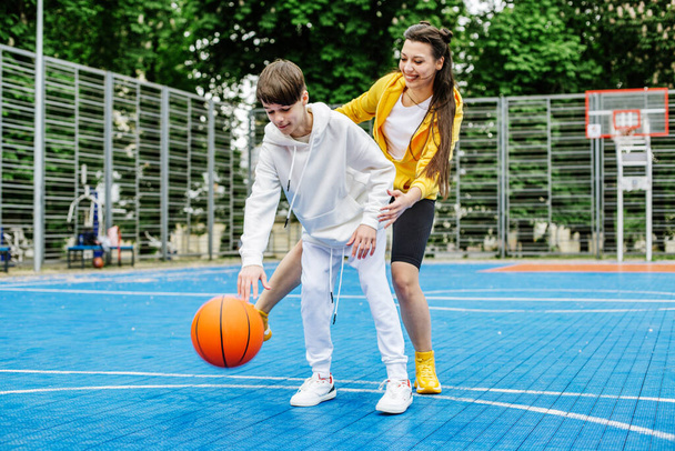 Chica y su hermano menor, adolescente, jugar al baloncesto en la cancha de baloncesto moderna bajo el cielo abierto. Concepto de deportes, aficiones y estilo de vida saludable. - Foto, Imagen
