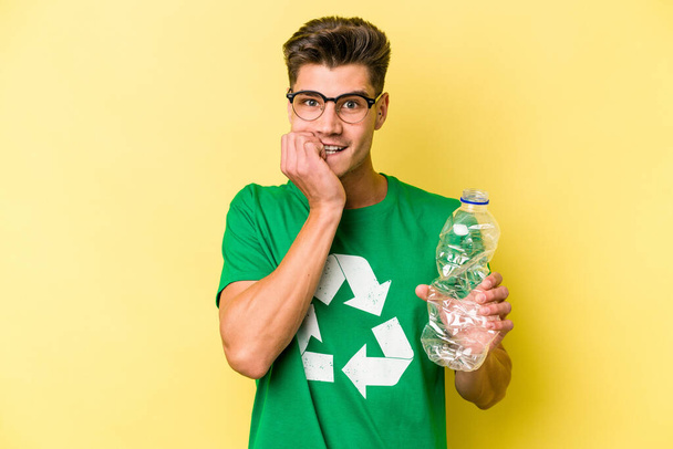 Joven hombre caucásico sosteniendo una botella de plástico para reciclar aislado sobre fondo amarillo mordiendo las uñas, nervioso y muy ansioso. - Foto, Imagen