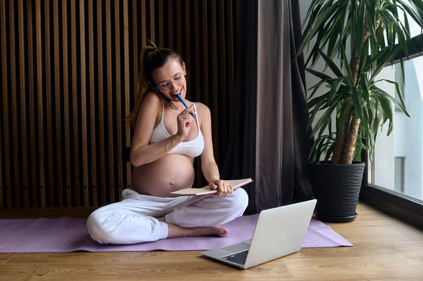 Gözlüklü güzel hamile bir iş kadını evde çalışan bir dizüstü bilgisayar ve not defteri kullanarak yerde oturuyor. - Fotoğraf, Görsel