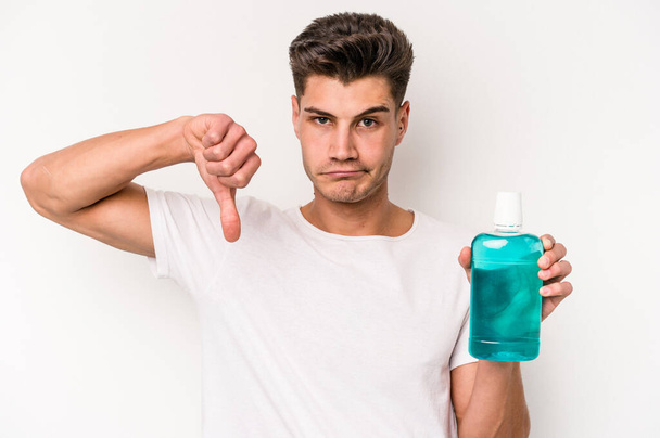 Young caucasian man holding mouthwash isolated on white background showing a dislike gesture, thumbs down. Disagreement concept. - Foto, afbeelding