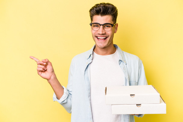 Young caucasian man holding pizzas isolated on yellow background smiling and pointing aside, showing something at blank space. - Foto, Imagem
