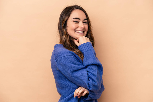 Young caucasian woman isolated on beige background smiling happy and confident, touching chin with hand. - Photo, image