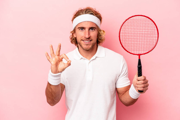 Jeune homme caucasien jouant au badminton isolé sur fond rose joyeux et confiant montrant ok geste. - Photo, image