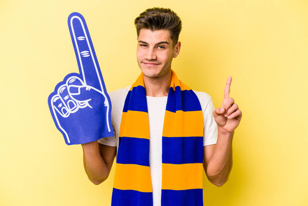 Fiatal sport fan man elszigetelt sárga háttér mutatja első számú ujját. - Fotó, kép