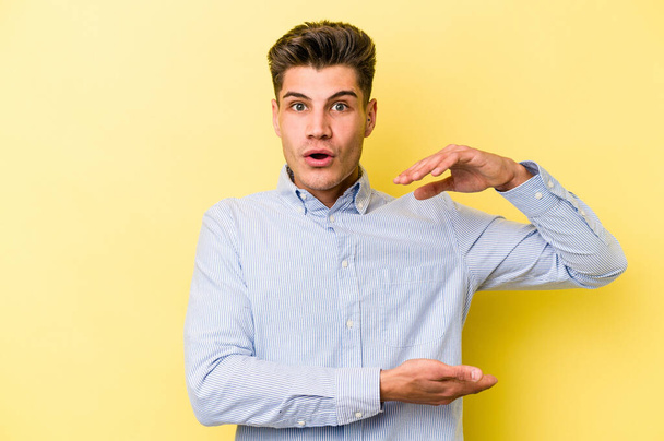 Young caucasian man isolated on yellow background holding something with both hands, product presentation. - Foto, Bild