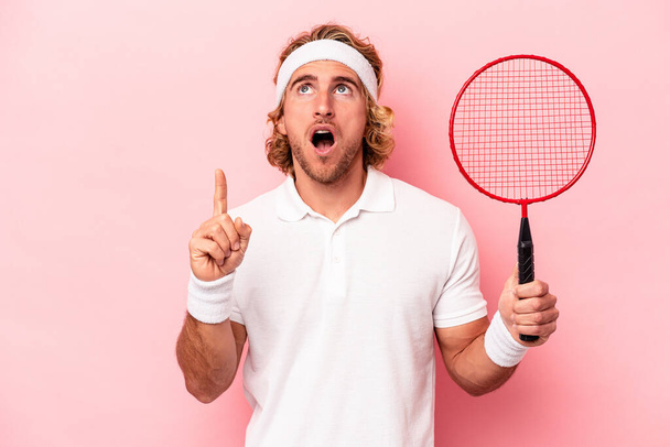 Young caucasian man playing badminton isolated on pink background pointing upside with opened mouth. - Foto, immagini