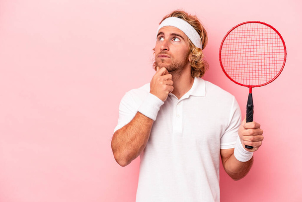 Young caucasian man playing badminton isolated on pink background looking sideways with doubtful and skeptical expression. - Fotoğraf, Görsel