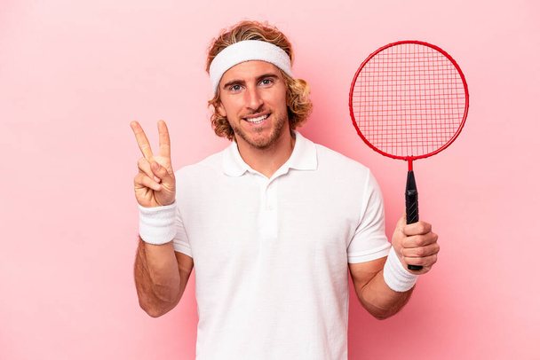 Young caucasian man playing badminton isolated on pink background joyful and carefree showing a peace symbol with fingers. - Photo, Image