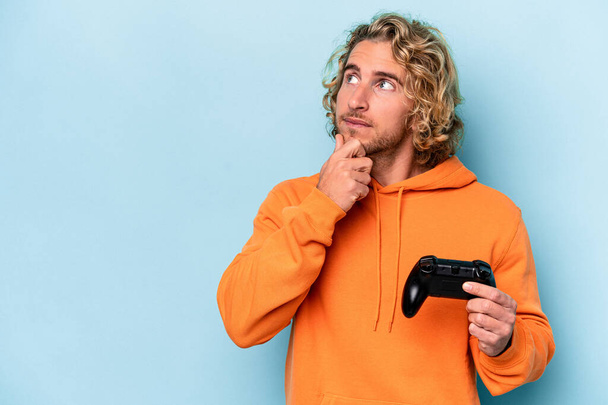 Young caucasian man playing with a video game controller isolated on blue background looking sideways with doubtful and skeptical expression. - Fotoğraf, Görsel