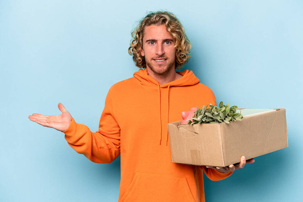 Young caucasian man making a move while picking up a box full of things isolated on blue background showing a copy space on a palm and holding another hand on waist. - Fotoğraf, Görsel